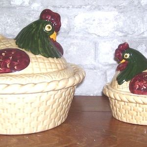 May include: Two ceramic hen-shaped containers. The lids are shaped like hens with green heads, red combs, and colorful tail feathers. The containers are cream-colored with a woven basket design. One is larger than the other.