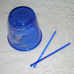 May include: A cobalt blue glass cup with an etched design of a windmill and waves. Two blue plastic stirrers are next to the cup.