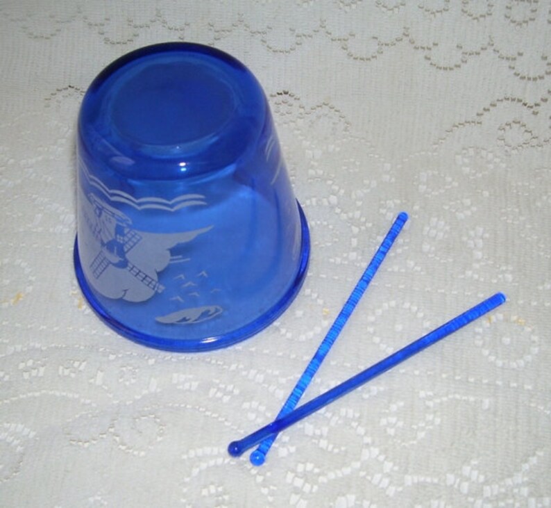 May include: A cobalt blue glass cup with an etched design of a windmill and waves. Two blue plastic stirrers are next to the cup.