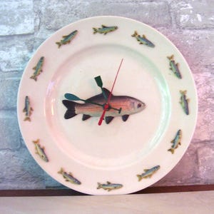 May include: A white ceramic plate repurposed as a clock, featuring a central fish design with a red and green clock hand. The plate is adorned with small fish illustrations around the rim, creating a nautical theme.