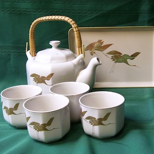 May include: A white ceramic tea set with a bamboo handle on the teapot and a tray. The set features a gold crane design. The set includes a teapot, four cups, and a tray.