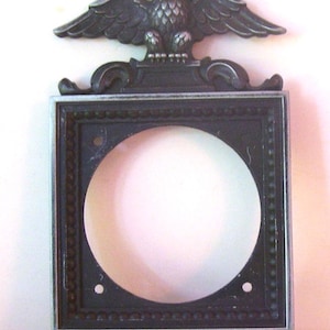 May include: A decorative metal frame with an eagle emblem at the top. The frame is square with a circular opening in the center. The eagle is detailed with outstretched wings, and the frame has an ornate design.
