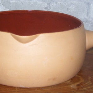 May include: A terracotta fondue pot with a brown interior and a handle. The pot is on a wooden surface.