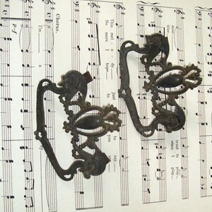May include: Two ornate metal drawer pulls with a decorative floral design. The pulls are made of a dark metal and have a vintage look.