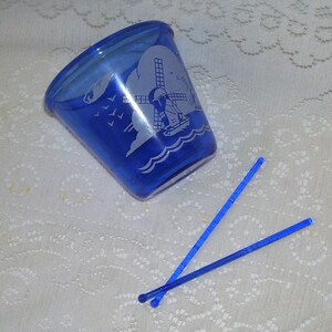 May include: A cobalt blue glass cup with a white windmill and boat design. Two blue plastic stirrers are next to the cup.
