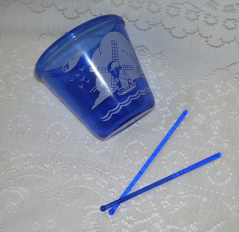May include: A cobalt blue glass cup with a white windmill and boat design. Two blue plastic stirrers are next to the cup.