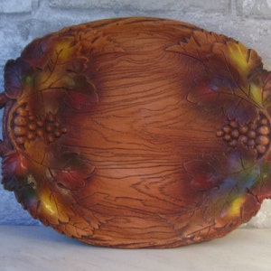 May include: A brown wooden serving tray with two handles and a carved design of grapes and leaves. The tray has a wood grain pattern and is painted with shades of brown, red, and yellow.