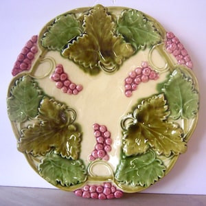 May include: A decorative ceramic plate with a grape and leaf design. The plate features green leaves, pink grapes, and a cream-colored center. The rim is scalloped, and the design is raised.