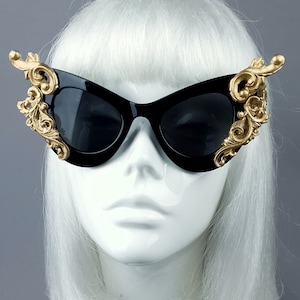 May include: Black cat-eye sunglasses with gold ornate detailing on the frames. The sunglasses are on a white mannequin head with white hair.
