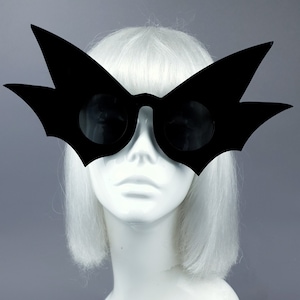 May include: A pair of black bat-shaped sunglasses on a white mannequin head with a white wig.