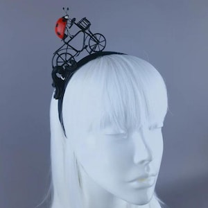 May include: A black headband with a red ladybug riding a black bicycle. The bicycle is made of wire and has a basket on the front.