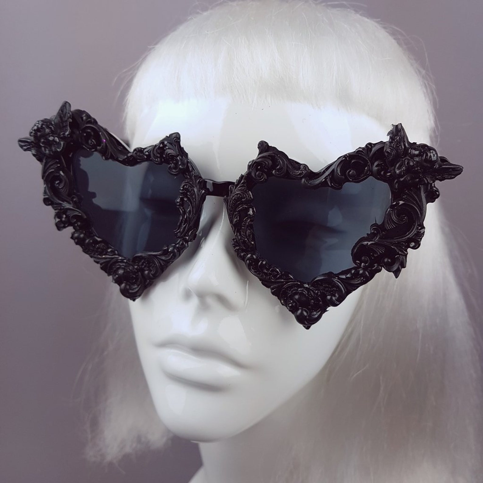 Heart-shaped gothic sunglasses with ornate black baroque frames and bat wing accents, displayed on white wig mannequin head