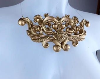 May include: A gold-colored, ornate, decorative necklace with a cherub design. The necklace is made of metal and has a baroque style.