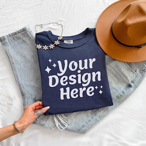 May include: A navy blue t-shirt with the text "Your Design Here" in white, paired with light blue jeans and a brown fedora hat. A silver necklace with flower accents is draped on the shirt.