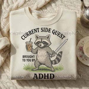 May include: Cream-coloured sweatshirt featuring a raccoon graphic holding a coffee and a sword. Text reads "CURRENT SIDE QUEST BROUGHT TO YOU BY ADHD". The raccoon stands on green grass.