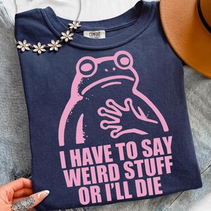 Könnte beinhalten: Ein marineblaues T-Shirt mit einer hellrosa Grafik eines Frosches, der seine Hand auf die Brust legt. Unter dem Frosch steht der Text: I HAVE TO SAY WEIRD STUFF OR I'LL DIE. Das T-Shirt hat ein Comfort Colors-Etikett am inneren Halsausschnitt.