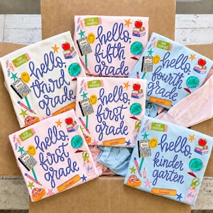 Pode incluir: Uma coleção de camisetas dobradas em branco, rosa claro e azul claro, cada uma com gráficos de temática escolar. Os designs incluem o texto hello third grade, hello fifth grade, hello fourth grade, hello first grade e hello kindergarten.