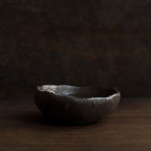 May include: A small, dark gray ceramic bowl with a rough, uneven, and textured surface. The bowl features a thick, organic rim with an irregular shape and a deep, rounded basin, showcasing a matte finish that appears weathered and hand-formed.
