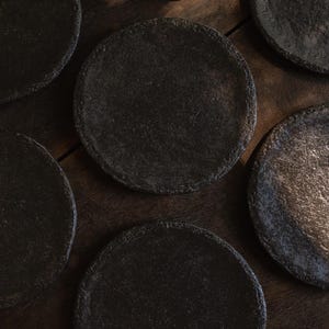 May include: A collection of circular plates with a dark, textured, matte finish. Each plate features a slightly uneven, rugged edge and a deep charcoal gray color, arranged in a cluster to display their individual surface variations and dark tones.