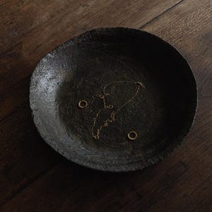 May include: A dark, textured, circular bowl with a rough, matte finish containing a delicate gold-toned chain necklace and two small gold-toned hoop earrings resting in the center of the basin.