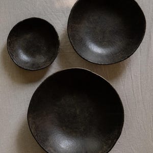 May include: Three circular bowls with a dark, textured, matte black finish. The set includes one small bowl and two larger bowls of similar size, all featuring irregular, organic edges and a deep, concave interior surface design.