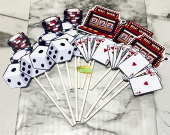 May include: A set of 15 casino-themed cupcake toppers. The toppers feature playing cards, dice, poker chips, and slot machines. The toppers are printed on white cardstock and attached to white sticks.
