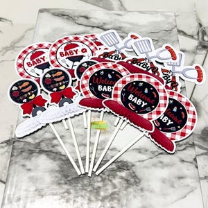 Könnte beinhalten: Ein Set mit 12 Cupcake-Toppern mit Grill-Thema, roten und weißen Karomustern, Grillwerkzeugen und dem Text "Baby Q" und "Willkommen Baby".