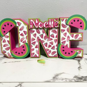 Puede incluir: Letras "UNO" rosas y verdes con un patrón de sandía y el nombre "Noelle" en letras rosas.