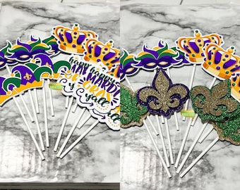 May include: A set of 12 Mardi Gras themed cupcake toppers. The toppers feature a variety of designs, including crowns, masks, fleur-de-lis, and a sign that says "Happy Mardi Gras y'all".