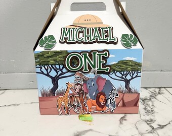 May include: A white favor box with a safari theme. The box has a handle and is decorated with a green jungle scene, a brown hat, and the words "Michael One".