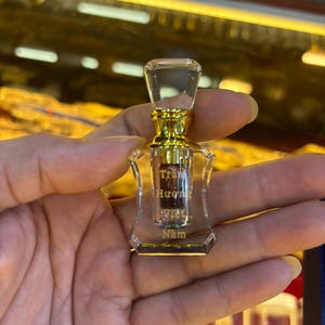 May include: A small, clear glass perfume bottle with a gold-colored cap and base. The bottle has a rectangular shape with a slightly curved design. The bottle contains a liquid and has text in a foreign language.