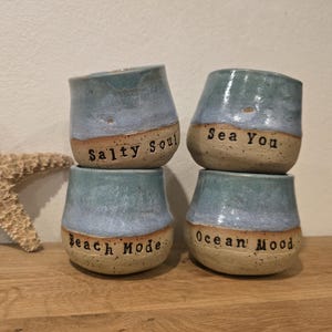 Könnte beinhalten: Vier gestapelte Keramikbecher, jeder mit einem einzigartigen Satz: "Salty Soul", "Sea You", "Beach Mode" und "Ocean Mood". Die Becher haben eine blau-grüne Glasur auf einer beigen Basis. Links ist ein Seestern.