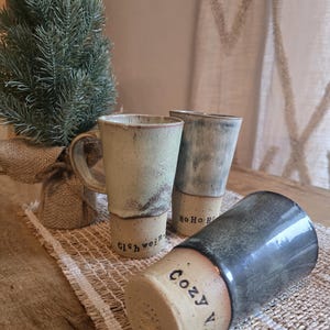Könnte beinhalten: Drei Keramikbecher mit rustikalem, handgefertigtem Aussehen. Die Becher sind in Beige-, Grau- und Blautönen gehalten und tragen die Aufschriften "Gluhwein", "Ho Ho Ho" und "Cozy Vibes". Im Hintergrund steht ein kleiner Nadelbaum.