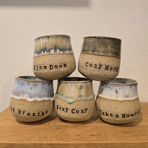 Könnte beinhalten: Fünf handgefertigte Keramikbecher mit gesprenkelter beigefarbener Basis und glasierten Oberteilen in Blau-, Grün- und Brauntönen. Jeder Becher hat einen anderen Schriftzug: "Slow Down", "Cozy Mood", "Just Breathe", "Stay Cozy" und "Take a Moment".