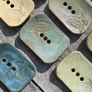 May include: Handmade ceramic soap dishes in shades of blue, green, and beige. Each dish has a raised turtle motif and three drainage holes. The rectangular dishes are displayed on a wooden surface.