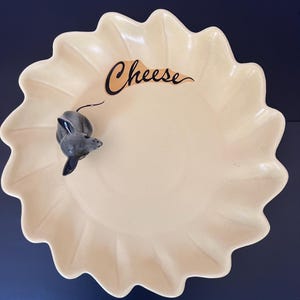 May include: A cream-colored ceramic serving dish with a scalloped edge and a glossy finish. The word Cheese is written in black cursive script on the surface. A small, three-dimensional gray mouse figurine is attached to the inner rim of the dish.