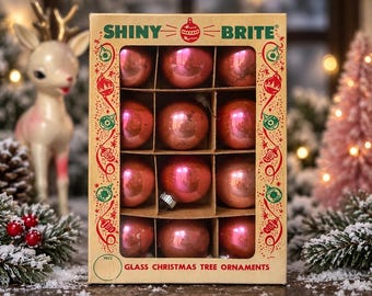 May include: A rectangular cardboard box containing twelve round, shiny pink glass Christmas tree ornaments. The box features the text SHINY BRITE at the top and GLASS CHRISTMAS TREE ORNAMENTS at the bottom, with decorative red and green illustrations.