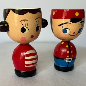 May include: Two wooden egg cups shaped like stylized figures. The left figure has dark hair, a red shirt with white horizontal stripes, and a black base. The right figure wears a red hat with yellow dots, a red jacket with yellow buttons, and a blue base.