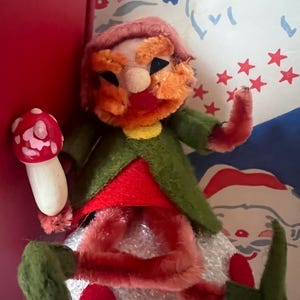 May include: A small elf figurine with a pink fabric cap, orange pipe cleaner hair, and a green felt jacket over a red tunic. The elf has pipe cleaner limbs, sits on a white textured sphere, and holds a small white and red mushroom ornament.