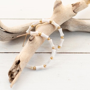 Puede incluir: Pendientes de aro blancos con detalles dorados. Los pendientes están hechos de pequeñas cuentas redondas que se ensartan para formar un círculo.