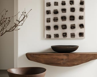 May include: A modern interior scene featuring a large, dark brown wooden bowl on the floor. A smaller, similar bowl sits on a floating wooden shelf. Above, a square artwork with a grid of dark, textured squares hangs on a white wall.