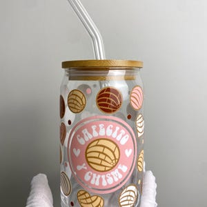 May include: A clear glass mug with a pink and white design featuring the text "Cafecito y Chisme" and a brown, white, and pink pastry design. The mug has a bamboo lid and a clear glass straw.