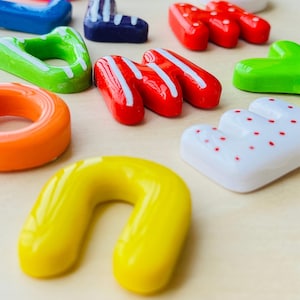 Puede incluir: Una colección de objetos brillantes en forma de letras, de colores vivos, incluyendo rojo, verde, amarillo y azul. Algunas letras tienen rayas blancas o puntos rojos. Las letras están dispuestas sobre una superficie clara.
