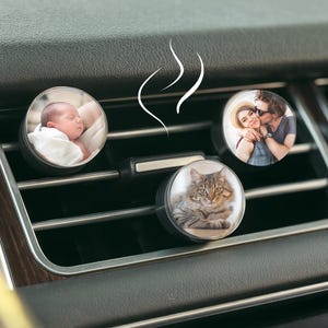 May include: Three round car air fresheners with personalized photos. One shows a sleeping baby, another a couple embracing, and the third a fluffy cat. The fresheners are black and sit in a car's air vent.
