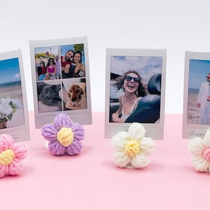May include: Four white photo frames with different photos inside, each with a crocheted flower in a different color: purple, white, pink, and light pink. The flowers are sitting on a pink surface.