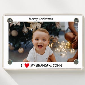 May include: A rectangular photo frame with a clear border and silver fasteners. The frame displays a photo of a smiling baby with the text "Merry Christmas" and "I ❤️ MY GRANDPA, JOHN".
