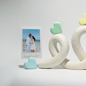 Könnte beinhalten: Weiße Keramikskulptur aus zwei ineinander verschlungenen Schleifen mit einem grünen, gelben und blauen Herzen auf jeder Schleife. Im Vordergrund befindet sich ein weißer Bilderrahmen mit einem Foto eines Paares in weißer Kleidung. Der Bilderrahmen trägt den Text "Happily Ever After".
