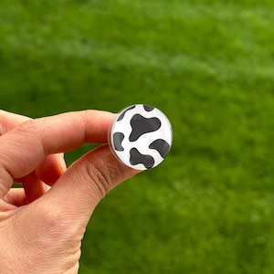 May include: A round, clear bottle stopper with a cow print design. The stopper features black irregular shapes on a white background. The stopper is held in a hand against a blurred green background.