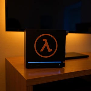 May include: A black, cube-shaped computer case featuring a prominent orange Greek lambda symbol enclosed within a circle on the front panel. A thin, horizontal blue LED light strip is positioned below the logo, with a small white power indicator light.