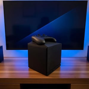 May include: A matte black, cube-shaped gaming console speaker with a textured front grille. A black wireless game controller with dual analog sticks and a central touchpad rests on the flat top surface of the speaker unit, positioned centrally.